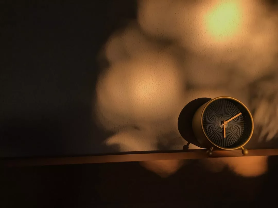 Eine runde GLAMESiA-Uhr mit goldenen Akzenten steht in einem Regal, beleuchtet von warmem Licht und umgeben von weichen Schatten an der Wand.
