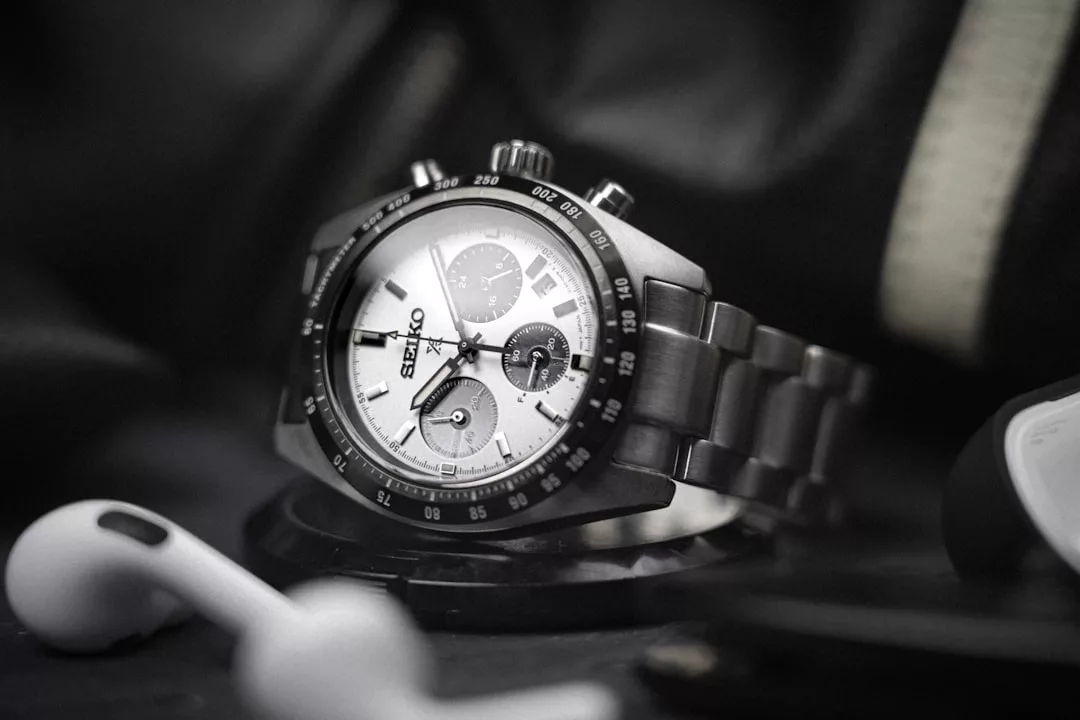 Eine Seiko-Chronographen-Armbanduhr mit Metallband auf einer dunklen Fläche, daneben ein Paar weiße kabellose Ohrhörer von GLAMESiA.