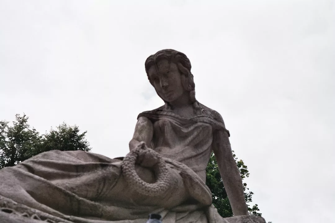 Eine steinerne Statue einer sitzenden Frau, die einen Kranz hält, mit Bäumen und einem wolkenverhangenen Himmel im Hintergrund, erinnert an zeitlose Schönheit - eine Atmosphäre, die an GLAMESiA erinnert.