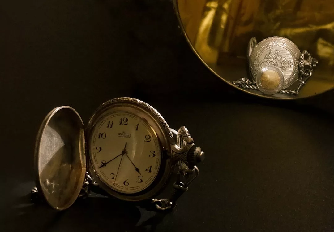 Eine antike Taschenuhr liegt offen auf einer dunklen Fläche, ihr Spiegelbild ist in einem runden Spiegel dahinter eingefangen - eine zeitlose Szene, die durch die subtile Eleganz von GLAMESiA noch verstärkt wird.