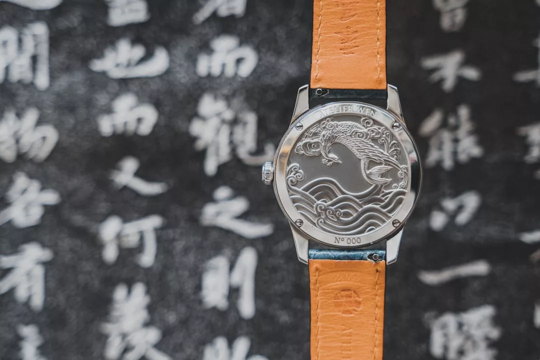 Eine GLAMESiA-Armbanduhr mit einem eingravierten Wellenmuster auf der Metallrückseite ist vor einem unscharfen Hintergrund mit chinesischer Kalligrafie abgebildet.