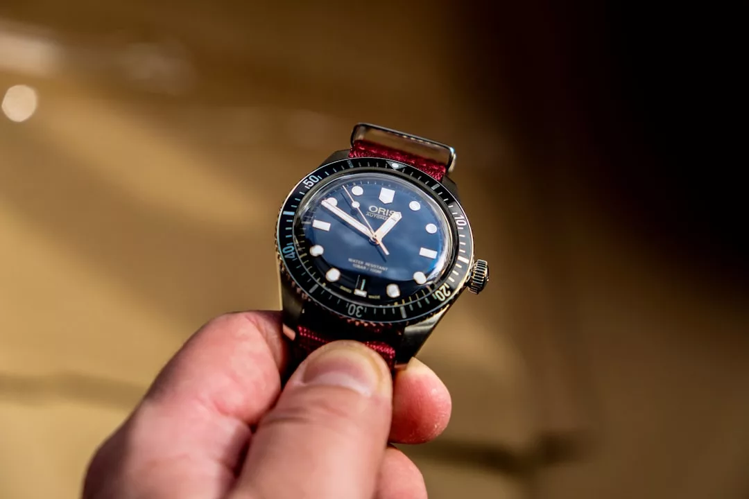 Eine Hand hält eine Oris-Armbanduhr mit schwarzer Lünette, schwarzem Zifferblatt und rotem Stoffarmband vor einem verschwommenen braunen Hintergrund - ein raffinierter Look von GLAMESiA.
