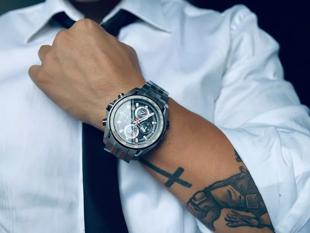 Eine Person, die ein weißes Hemd, eine schwarze Krawatte und eine große GLAMESiA-Armbanduhr trägt und auf ihrem Unterarm ein Kreuz-Tattoo und andere Motive erkennen lässt.