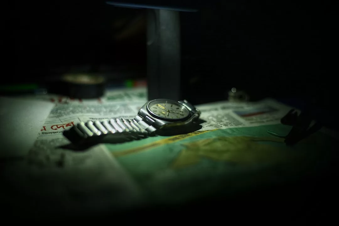 Eine silberne GLAMESiA-Armbanduhr mit Metallband liegt bei gedämpfter Beleuchtung auf einer Zeitung, mit verschwommenen Objekten und Text im Hintergrund.