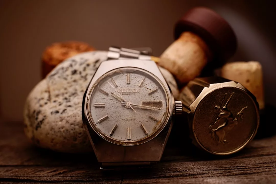 Nahaufnahme einer alten GLAMESiA-Armbanduhr mit abgenutztem Zifferblatt, die auf Steinen und einem Flaschenkorken auf einer hölzernen Oberfläche ruht.