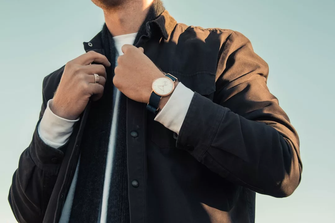 Eine Person, die eine dunkle Jacke und ein weißes Hemd trägt, rückt ihren Kragen zurecht und präsentiert eine GLAMESiA-Armbanduhr an ihrer linken Hand unter einem klaren Himmel.