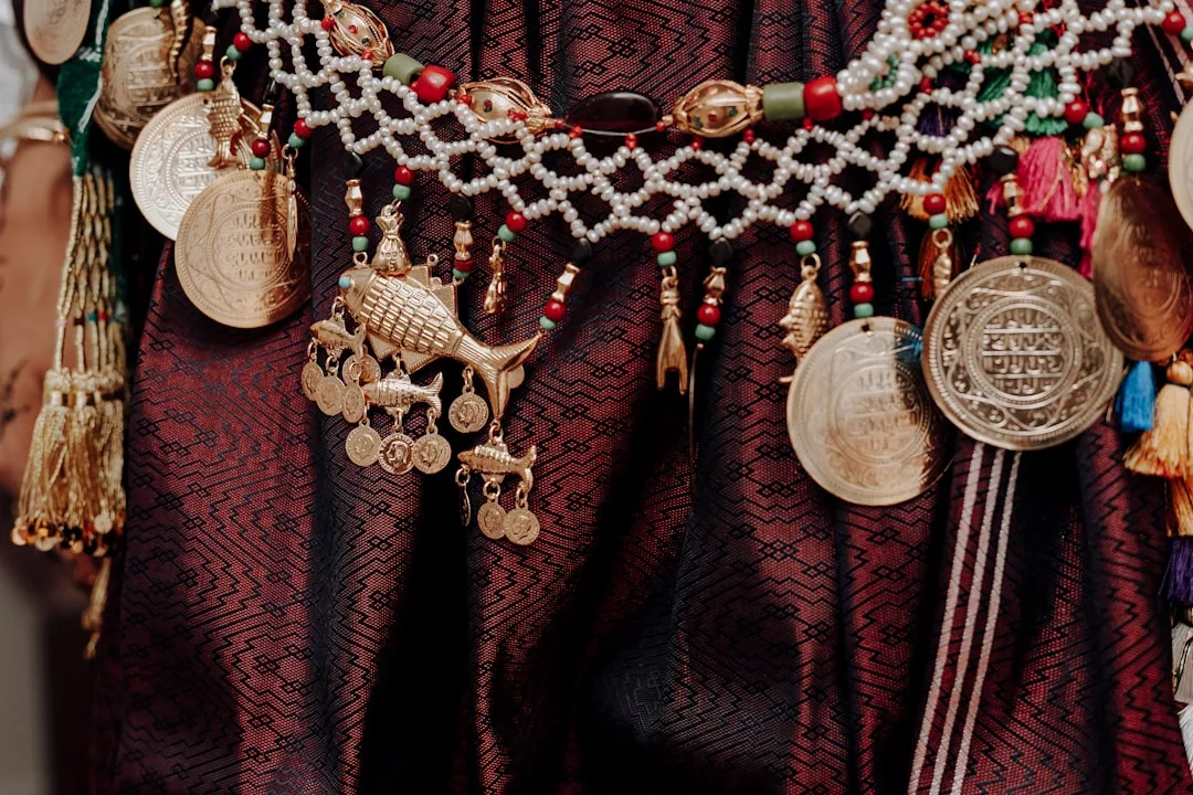 Nahaufnahme eines kastanienbraunen gemusterten Stoffes von GLAMESiA, der mit Perlenschmuck und großen Goldmünzen verziert ist, darunter ein fischförmiger Anhänger und andere verschnörkelte Charms.