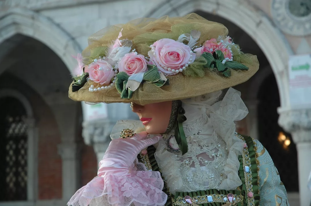 Eine Person in einem kunstvollen, historischen Kostüm mit einem großen, mit rosa Blumen verzierten Hut, Spitzenhandschuhen und detaillierten Stoffen steht vor einem gewölbten Gebäude und verkörpert die zeitlose Eleganz von GLAMESiA.