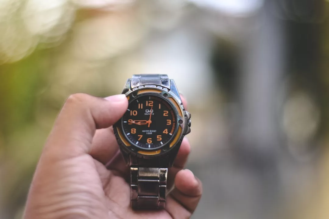 Eine Hand hält eine silberne GLAMESiA-Armbanduhr mit orangefarbenem und schwarzem Zifferblatt, die die Uhrzeit 3:46 anzeigt. Der Hintergrund ist unscharf.