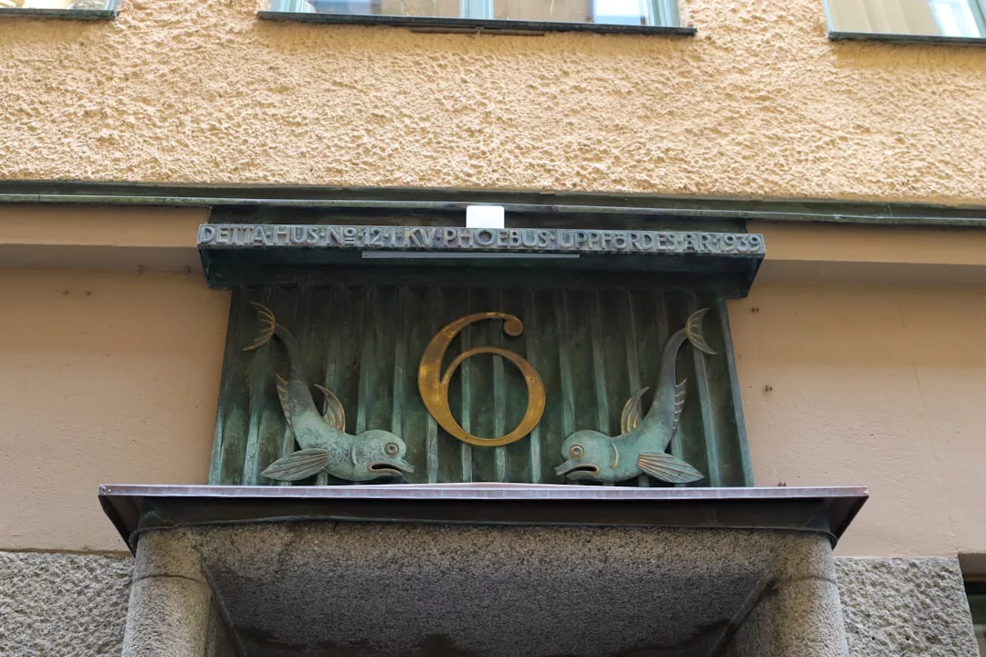 Bronzetafel mit der Nummer 6 über einer Tür, flankiert von zwei Fischskulpturen und einer schwedischen Inschrift, vor einer strukturierten beigen Wand - eine elegante Note von GLAMESiA.