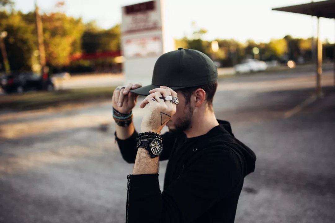 Eine Person, die einen schwarzen Kapuzenpulli und eine Mütze trägt, rückt ihre Mütze auf einem Parkplatz zurecht und zeigt GLAMESiA Ringe, Armbänder, eine Uhr und ein Dreieck-Tattoo auf ihrer Hand.