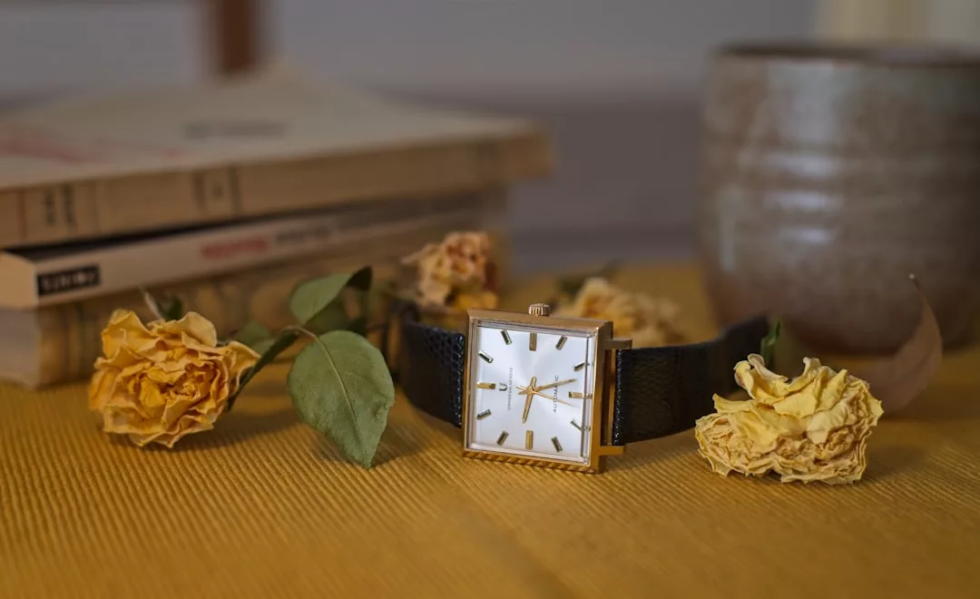 Eine quadratische GLAMESiA-Armbanduhr mit schwarzem Band ruht auf einer gelben Fläche, umgeben von getrockneten gelben Rosen, Büchern und einer Keramiktasse im Hintergrund.