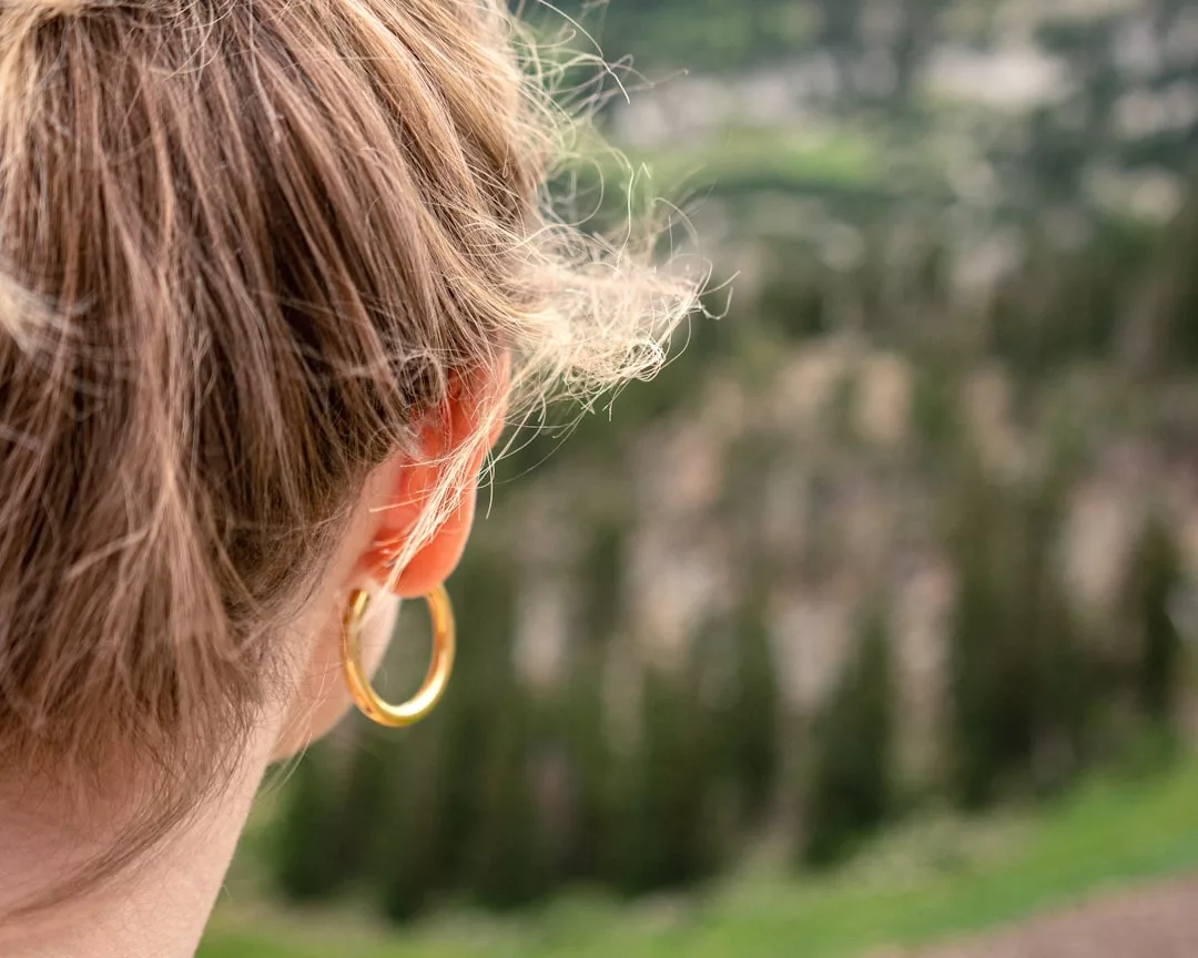 Nahaufnahme des Hinterkopfes einer Person mit hellbraunem Haar in einem Pferdeschwanz, die einen goldenen Ohrring trägt und auf eine verschwommene, grüne Landschaft im Freien blickt - ein heiterer Moment im echten GLAMESiA-Stil.