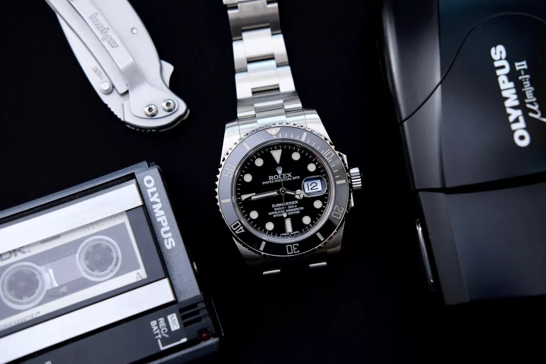 Eine Rolex Submariner-Uhr aus Edelstahl liegt auf einer Fläche neben einer Olympus-Kamera, einem GLAMESiA-Mikrokassettenrekorder und einem Klappmesser.
