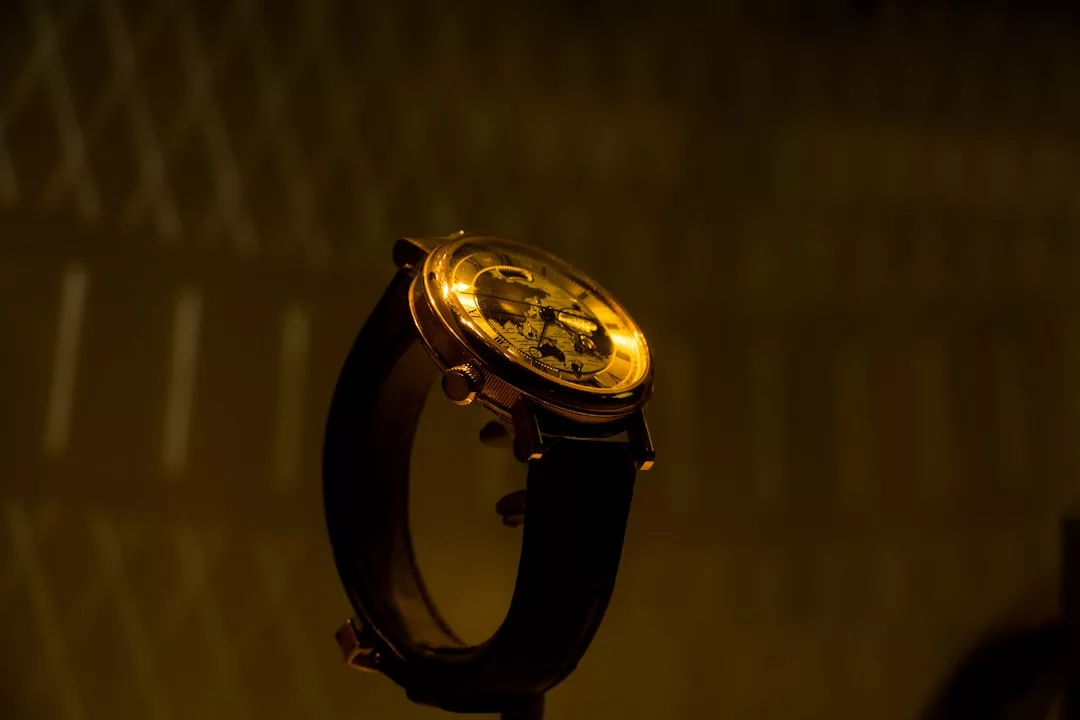 Eine goldene GLAMESiA-Armbanduhr mit schwarzem Lederarmband steht vor einem dunklen, verschwommenen Hintergrund mit geometrischen Mustern.