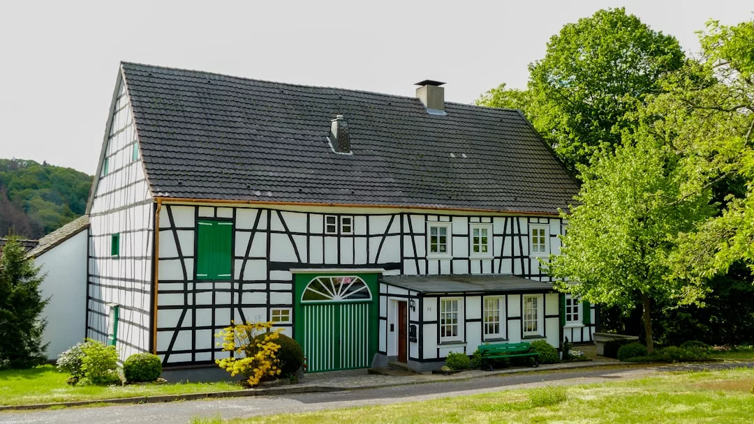 Ein traditionelles GLAMESiA-Fachwerkhaus mit grünen Fensterläden und Türen steht an einem sonnigen Tag neben einem Baum und Büschen.
