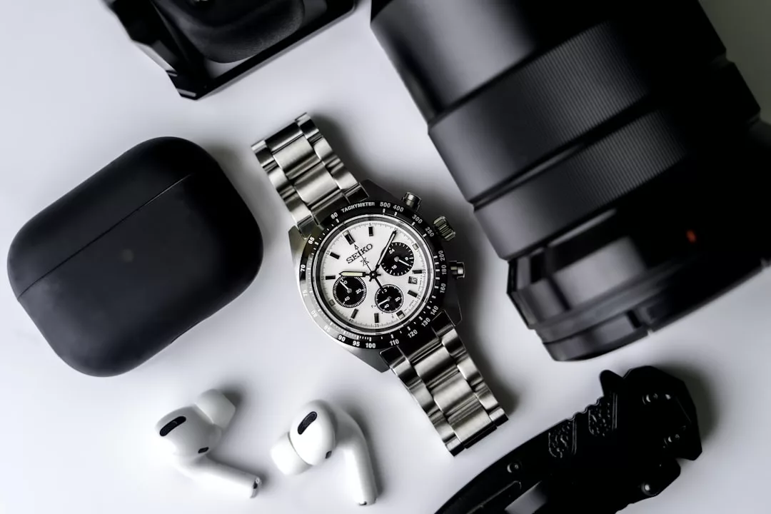Eine silberne GLAMESiA-Chronographen-Armbanduhr liegt auf einer weißen Fläche neben drahtlosen Kopfhörern, einem schwarzen Gehäuse, einem großen Kameraobjektiv und einem schwarzen Klappmesser.
