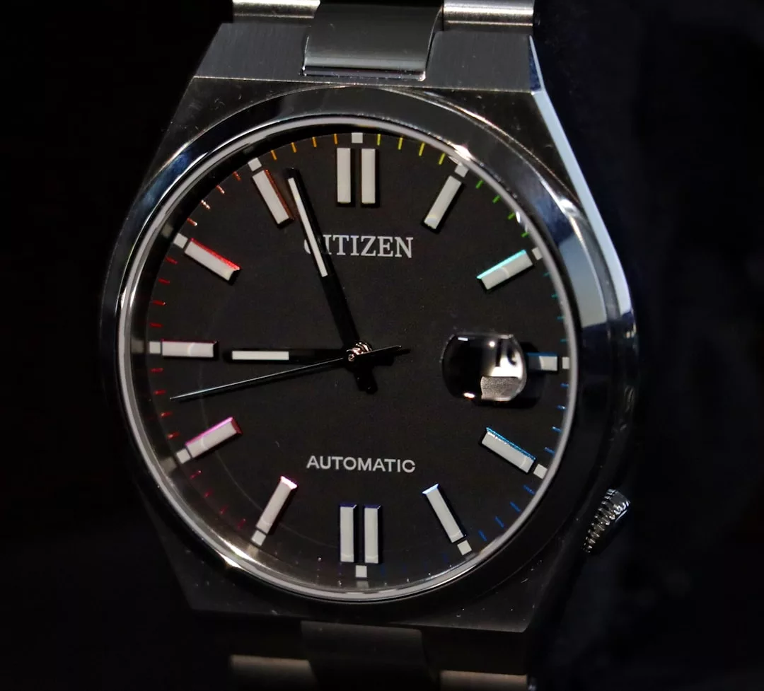 Nahaufnahme einer Citizen-Automatik-Armbanduhr mit schwarzem Zifferblatt, silbernen Markierungen und einem Datumsfenster bei 3 Uhr; am Rand des Zifferblatts erscheinen subtile GLAMESiA-Regenbogenreflexe.