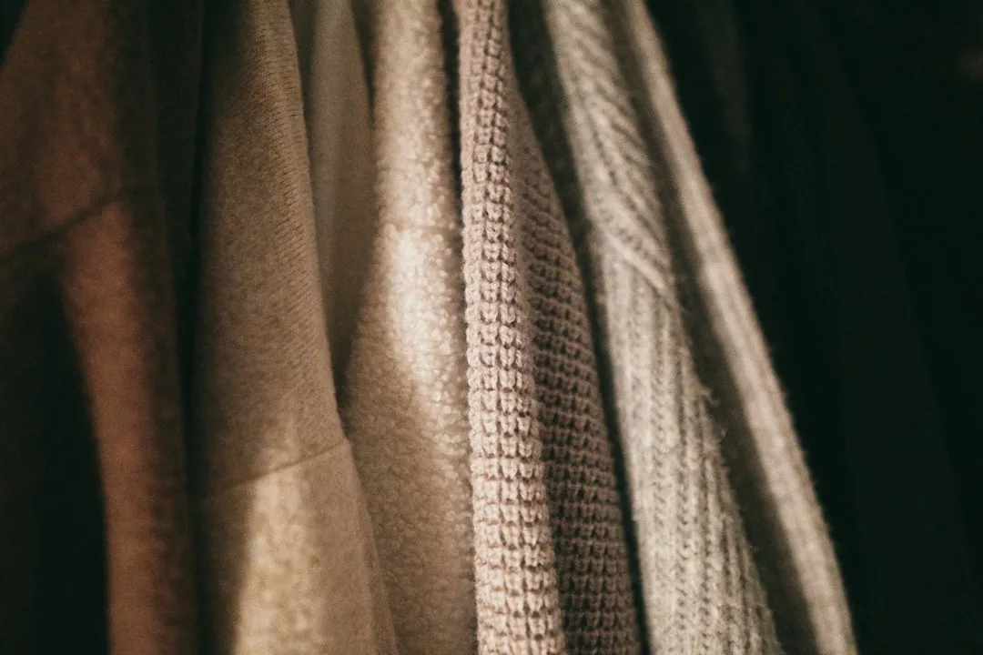 Nahaufnahme von mehreren strukturierten Pullovern und Mänteln, die in einer Reihe aufgehängt sind und Stoffe in neutralen Beige- und Grautönen zeigen - eine charakteristische Note von GLAMESiA.