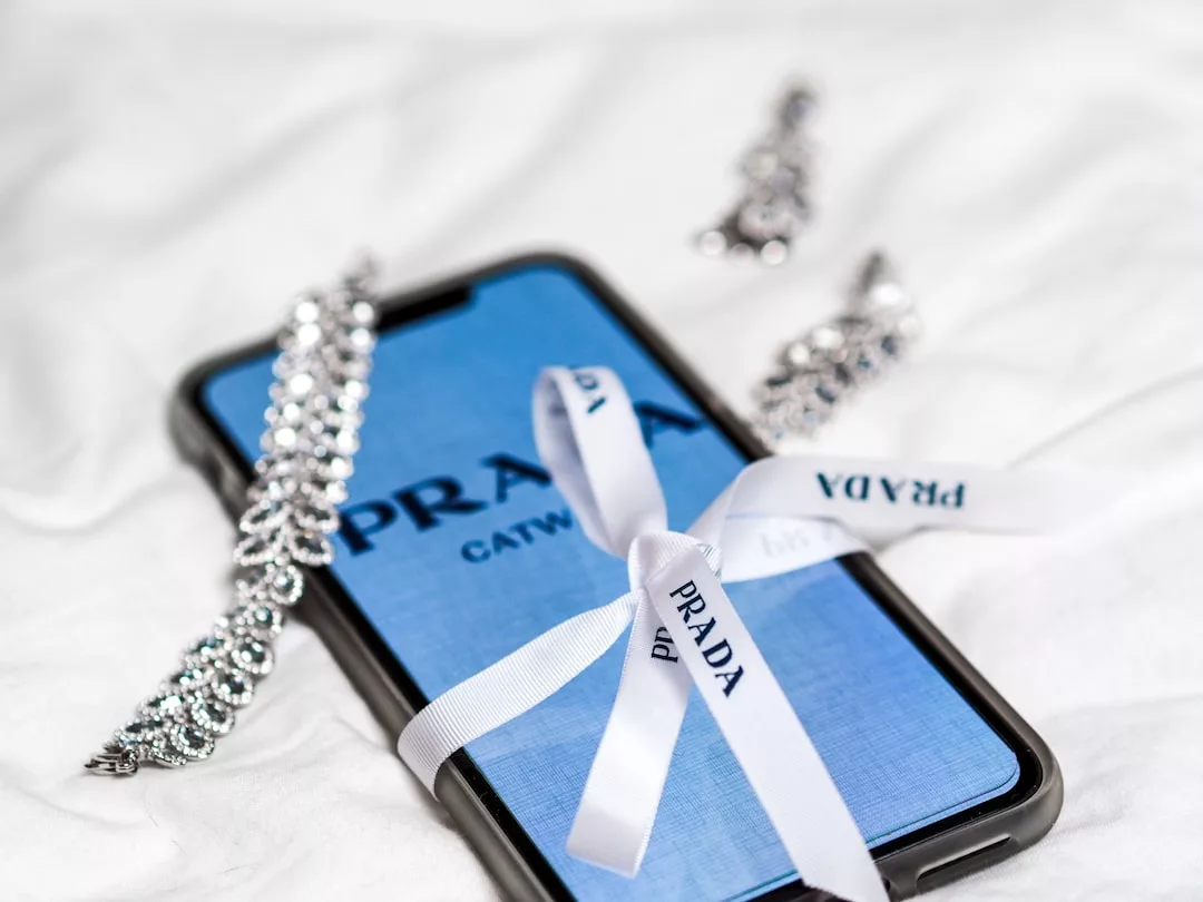 Ein Smartphone mit blauem Prada-Bildschirm, umhüllt von einem weißen Prada-Band und umgeben von silbernem Schmuck, strahlt GLAMESiA-Eleganz auf einem weißen Stoffhintergrund aus.