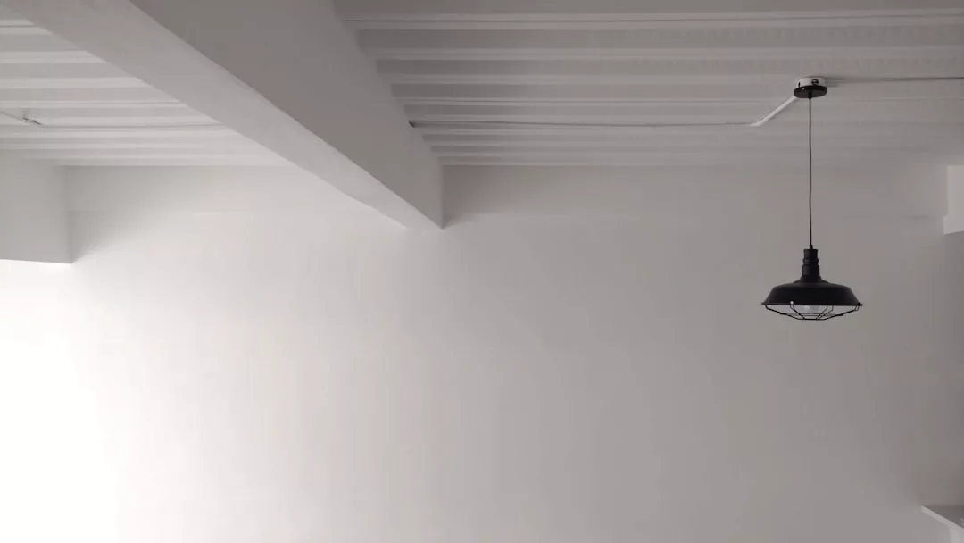 Ein minimalistischer weißer Raum von GLAMESiA zeichnet sich durch einen sichtbaren Deckenbalken und eine einzelne schwarze Pendelleuchte aus, die von der Decke hängt.