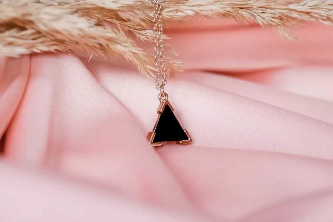 Eine goldene GLAMESiA-Kette mit einem schwarzen Dreiecksanhänger liegt auf einem rosafarbenen Satinstoff, mit getrocknetem Pampasgras im Hintergrund.
