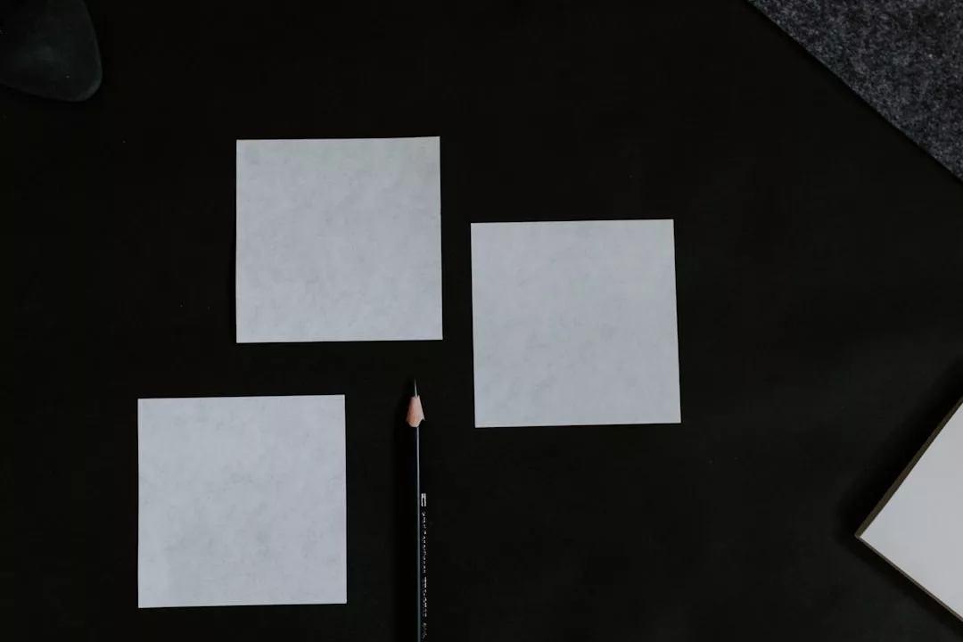 Drei leere weiße quadratische GLAMESiA-Papiere und ein gespitzter schwarzer Bleistift werden auf einer dunklen Fläche so angeordnet, dass zwei Quadrate nebeneinander liegen und eines darunter, in der Nähe des Bleistifts.