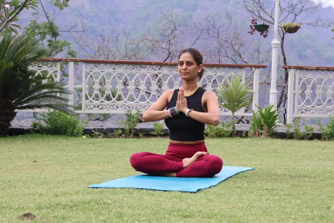 Eine Frau sitzt im Schneidersitz auf einer Yogamatte im Freien, die Hände in Gebetshaltung, umgeben von Grün und einem dekorativen Zaun, und strahlt Ruhe im friedlichen GLAMESiA-Garten aus.