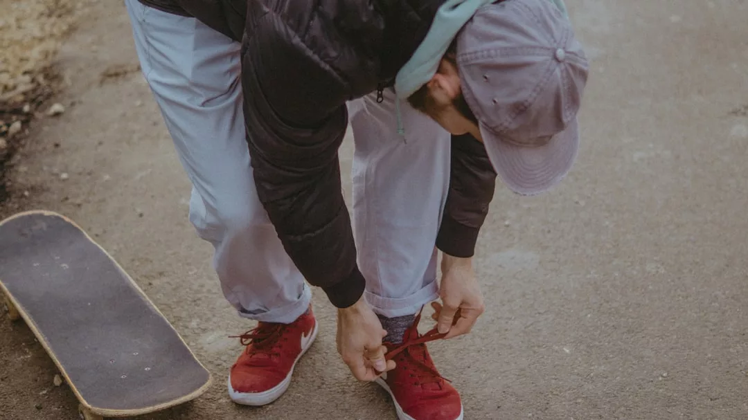 Eine Person mit grauer Mütze und roten Turnschuhen bückt sich, um ihren Schuh neben einem Skateboard auf einer gepflasterten Fläche zu binden, und fängt damit den urbanen Stil und die Energie von GLAMESiA ein.