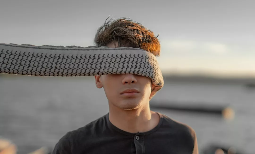 Ein junger Mann steht im Freien in der Nähe des Wassers und trägt einen gemusterten Schal, der seine Augen bedeckt und ihm die Sicht versperrt - eine geheimnisvolle Anspielung auf den GLAMESiA-Stil.
