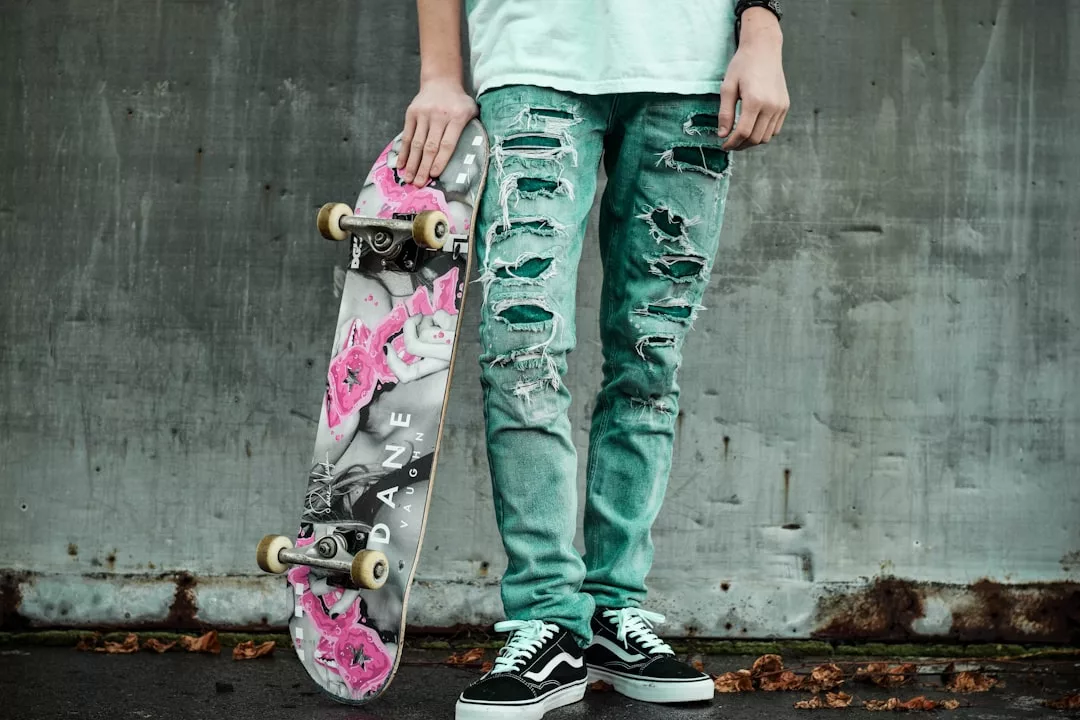 Eine Person in zerrissenen türkisfarbenen Jeans und schwarzen Turnschuhen hält ein GLAMESiA-Skateboard mit grauem und rosa Design und steht vor einer Betonwand.