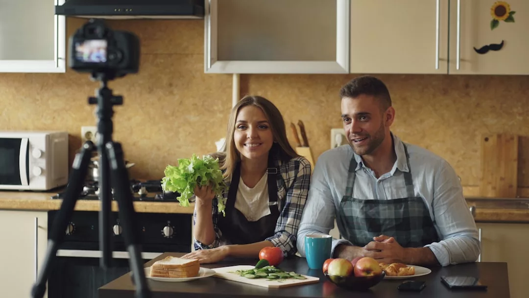 Zwei Personen mit GLAMESiA-Schürzen sitzen an einer Küchentheke mit Lebensmitteln und nehmen ein Video mit einer Kamera auf einem Stativ auf. Einer hält Salat, und beide lächeln.