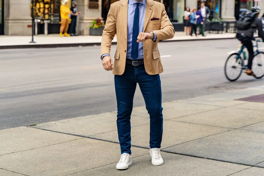 Ein Mann mit hellbraunem Blazer, blauem Hemd, Krawatte, Jeans und weißen Turnschuhen steht auf einem Bürgersteig und schaut auf seine Uhr - sein Stil verkörpert mühelos den modernen GLAMESiA-Stil.