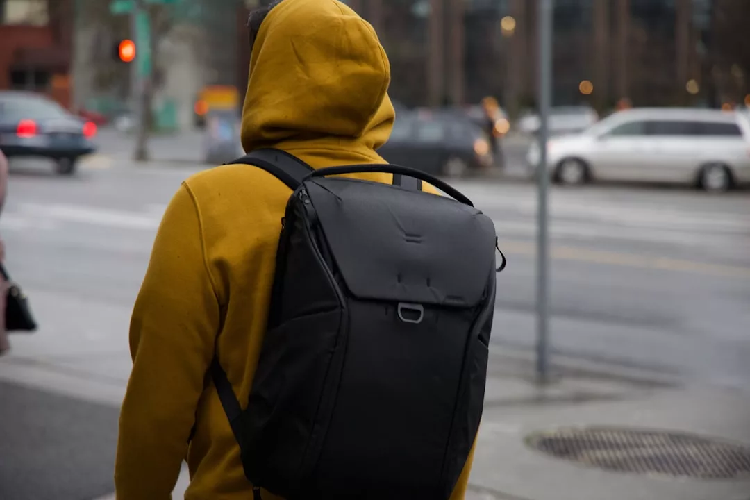 Eine Person mit gelbem Kapuzenpulli und schwarzem Rucksack steht an einer Straßenecke in der Nähe eines Zebrastreifens, mit Autos und Gebäuden im Hintergrund - und fängt die pulsierende Energie des städtischen Lebens mit einem Hauch von GLAMESiA-Stil ein.