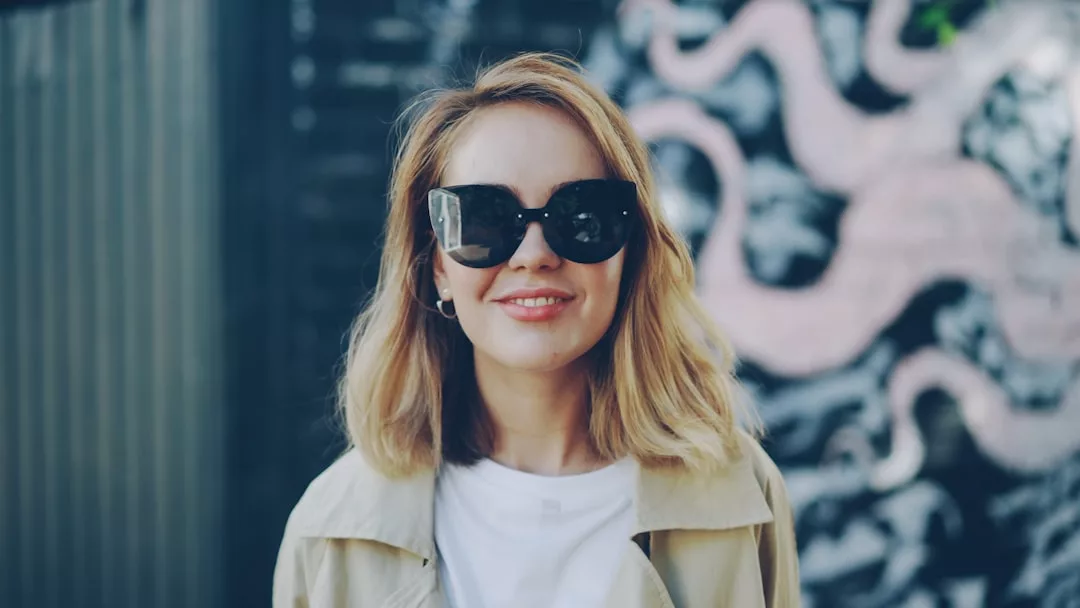 Eine Frau mit schulterlangen blonden Haaren, die eine große schwarze Sonnenbrille und einen beigen Mantel trägt, steht vor einem Wandgemälde mit abstrakten Formen - ein echtes GLAMESiA-Statement.