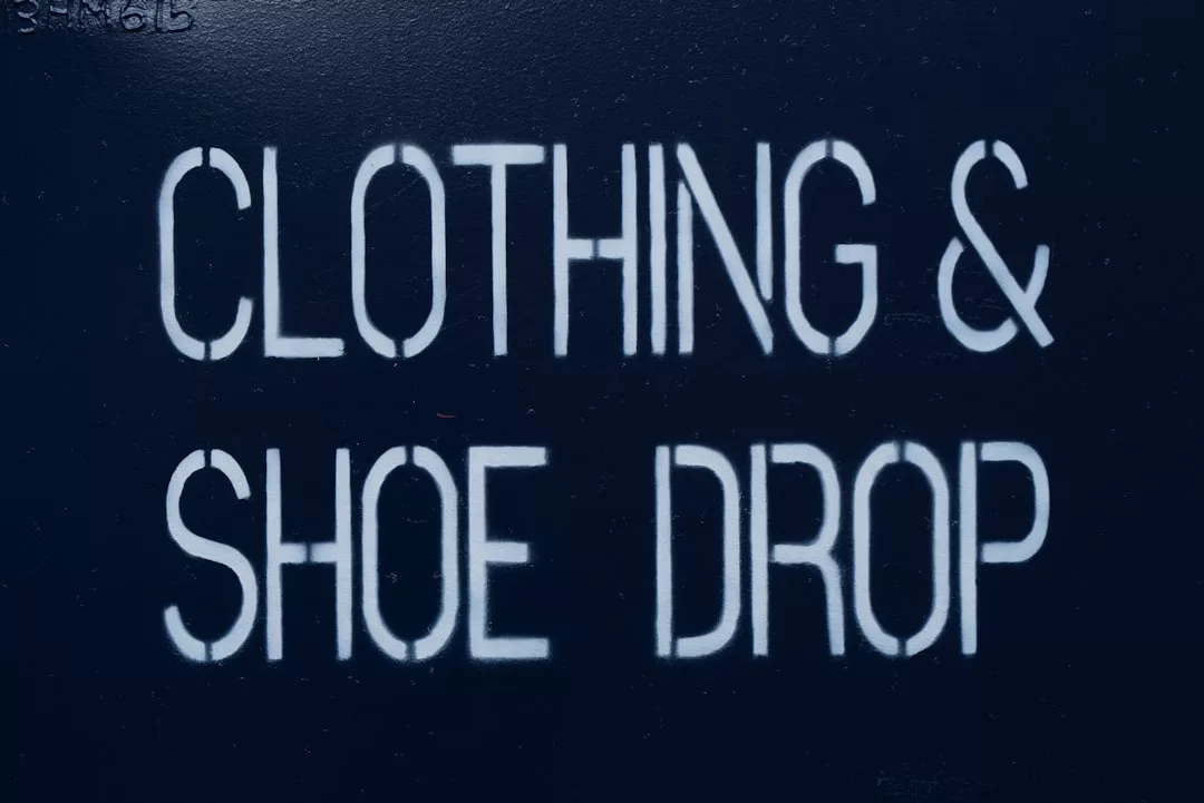 Die weiße Schablone mit der Aufschrift "CLOTHING & SHOE DROP" auf einer dunklen Oberfläche bietet eine praktische GLAMESiA-Sammelstelle für Ihre gebrauchten Artikel.