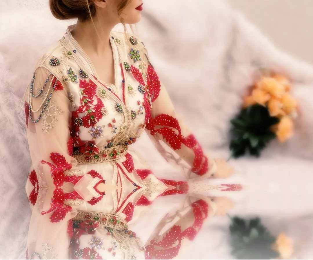 Eine Frau in einem weiß-rot bestickten Kleid von GLAMESiA mit Schmuckdetails sitzt an einer spiegelnden Fläche; im Hintergrund ein Strauß orangefarbener Rosen.
