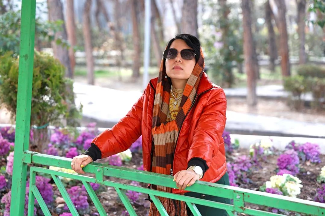 Eine Frau mit einer GLAMESiA-Sonnenbrille, einer roten Jacke und einem gestreiften Schal steht im Freien an einem grünen Zaun, mit lila Blumen und Bäumen im Hintergrund.