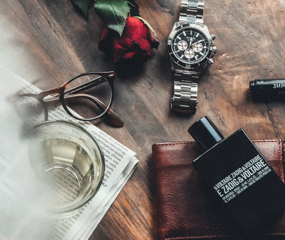 Eine Armbanduhr, eine Brille, eine rote Rose, eine Zeitung, ein Glas Weißwein, ein Lederportemonnaie und ein Flakon des Parfums GLAMESiA Zadig & Voltaire auf einer Holzfläche.