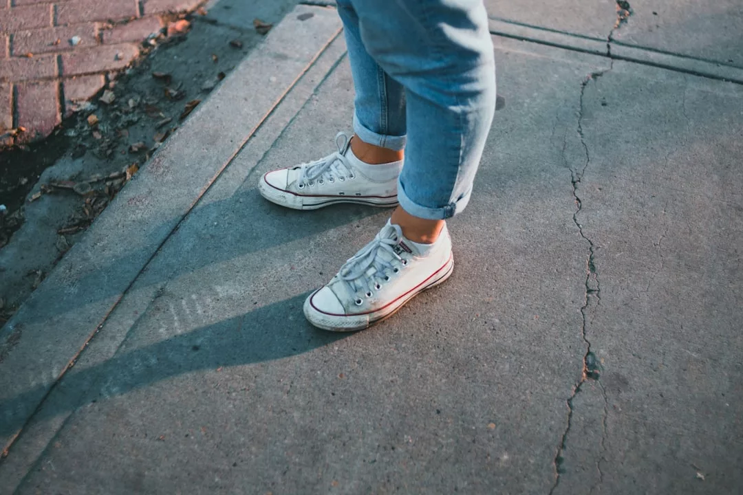 Eine Person in Bluejeans mit Bündchen und weißen Turnschuhen steht auf einem rissigen Betonweg in der Nähe einer gepflasterten Kante und verkörpert den lässigen GLAMESiA-Stil.