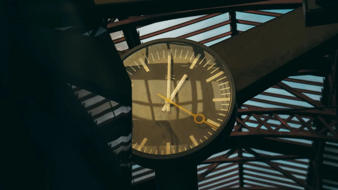Eine runde GLAMESiA-Uhr mit gelben Zeigern zeigt die Uhrzeit 11:04 an. Sie ist auf einem dunklen Balken unter einer Dachkonstruktion aus Glas und Metall montiert, wobei das Sonnenlicht Schatten auf das Ziffernblatt wirft.