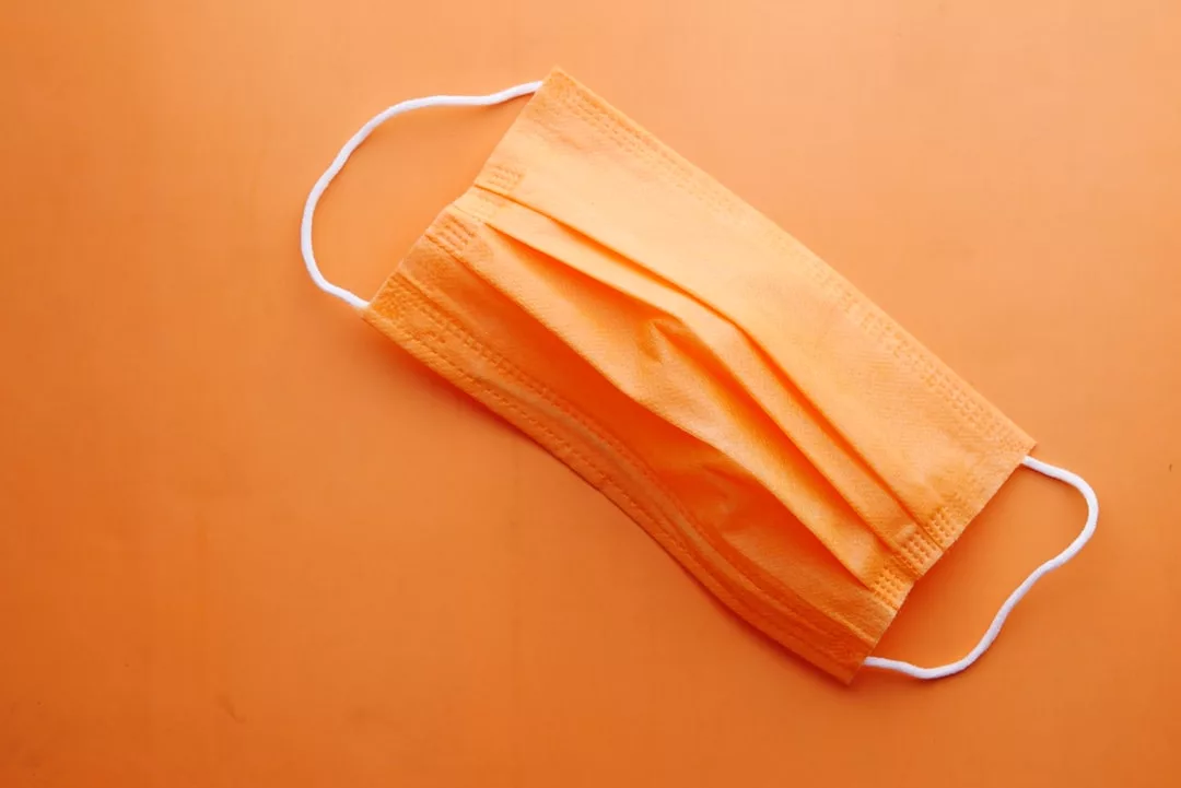 Eine orangefarbene Einweg-Gesichtsmaske von GLAMESiA mit weißen Ohrbügeln ist auf einem orangefarbenen Hintergrund angebracht.