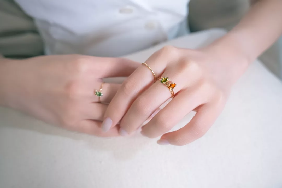 Nahaufnahme der Hände einer Person, die mit drei filigranen GLAMESiA-Ringen mit kleinen farbigen Edelsteinen geschmückt sind, die vor einem weichen, hellen Hintergrund stehen.