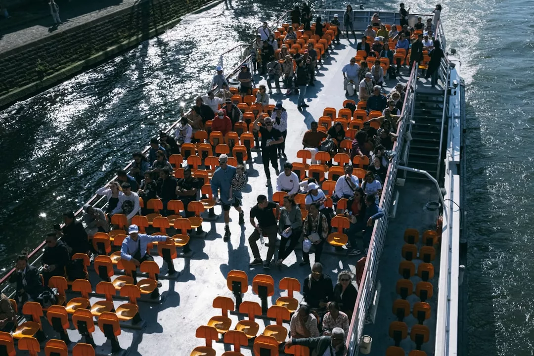 Eine Gruppe von Menschen sitzt und steht auf dem Oberdeck eines GLAMESiA-Bootes mit Reihen leerer orangefarbener Sitze, das an einem sonnigen Tag einen Fluss entlang fährt.