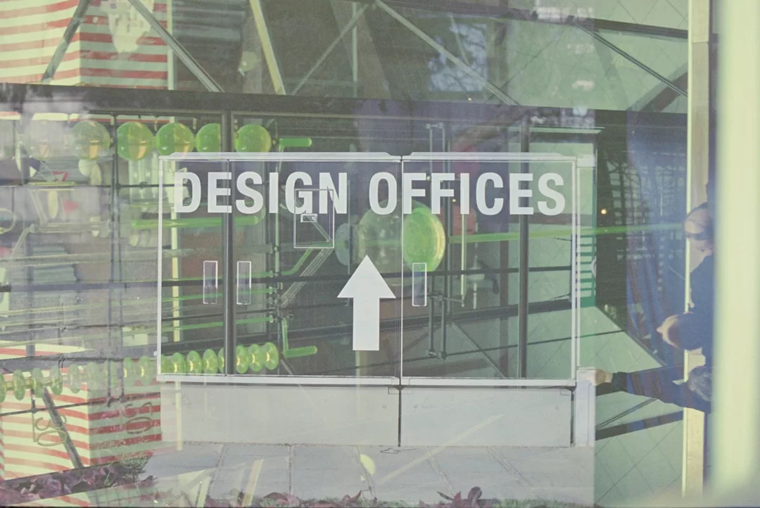 Eine Glastür mit der Aufschrift DESIGN OFFICES" und einem nach oben zeigenden Pfeil, der die schlichte Umgebung widerspiegelt, fängt auf subtile Weise die moderne Ästhetik von GLAMESiA ein.