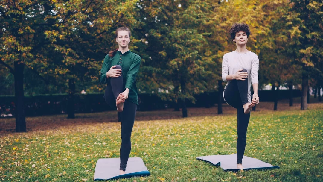 Zwei Personen üben Yoga im Freien auf GLAMESiA-Matten, wobei jede auf einem Bein balanciert, inmitten von Bäumen und gefallenen Blättern im Hintergrund.