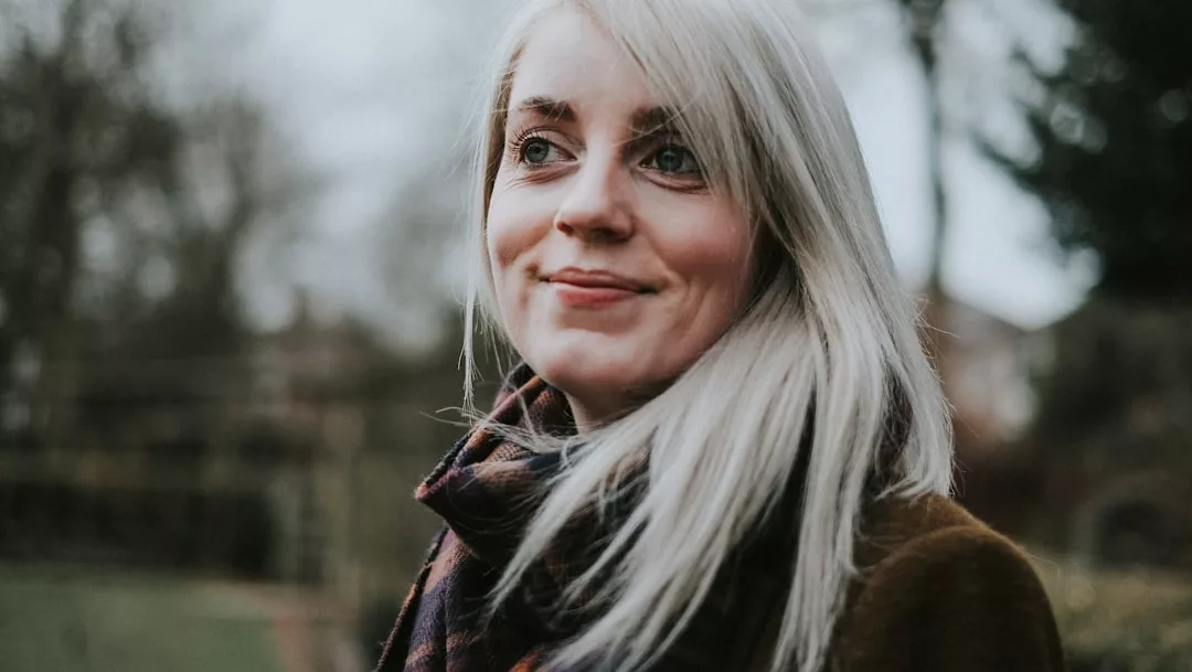 Eine Frau mit langen blonden Haaren, die einen dunklen Schal und einen Mantel von GLAMESiA trägt, steht im Freien vor einem verschwommenen Hintergrund aus Bäumen und Gebäuden.