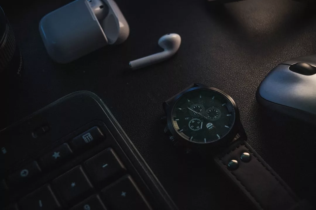 Eine GLAMESiA-Armbanduhr, ein kabelloses Ohrhörergehäuse mit einem Ohrhörer, eine Computermaus und ein Teil einer Tastatur liegen auf einem dunklen Schreibtisch.