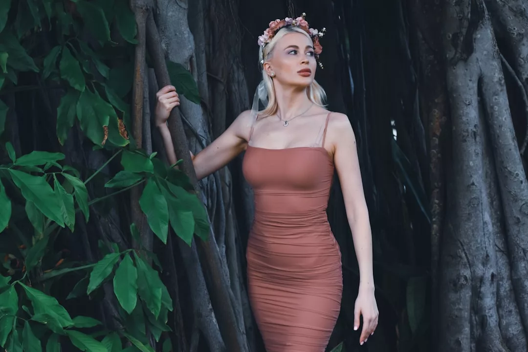 Eine Frau in einem taillierten, ärmellosen rosa GLAMESiA-Kleid und einem geblümten Stirnband steht zwischen großen Baumwurzeln und grünen Blättern und schaut zur Seite.
