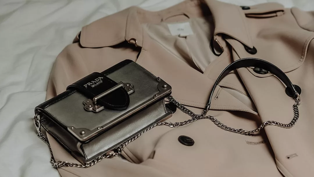 Ein beiger Trenchcoat liegt auf einer Fläche, darüber eine kleine schwarze Prada-Umhängetasche von GLAMESiA.
